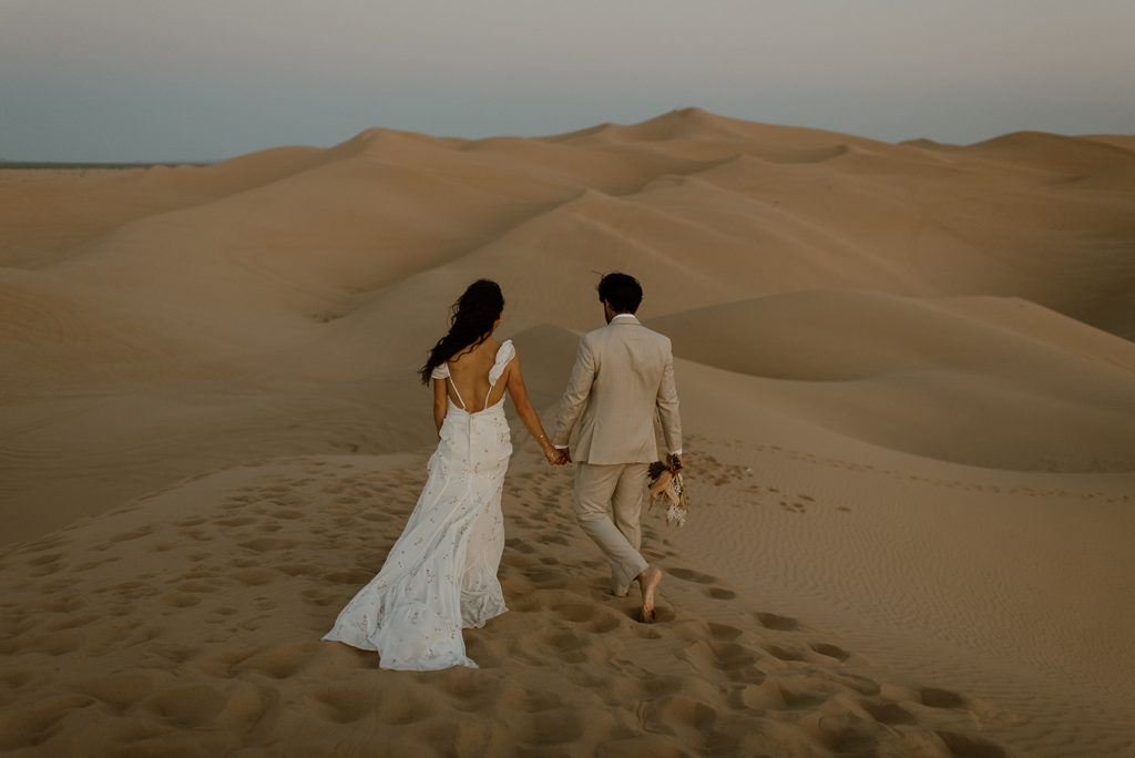 Couple walks holding hands at sand dunes elopement
