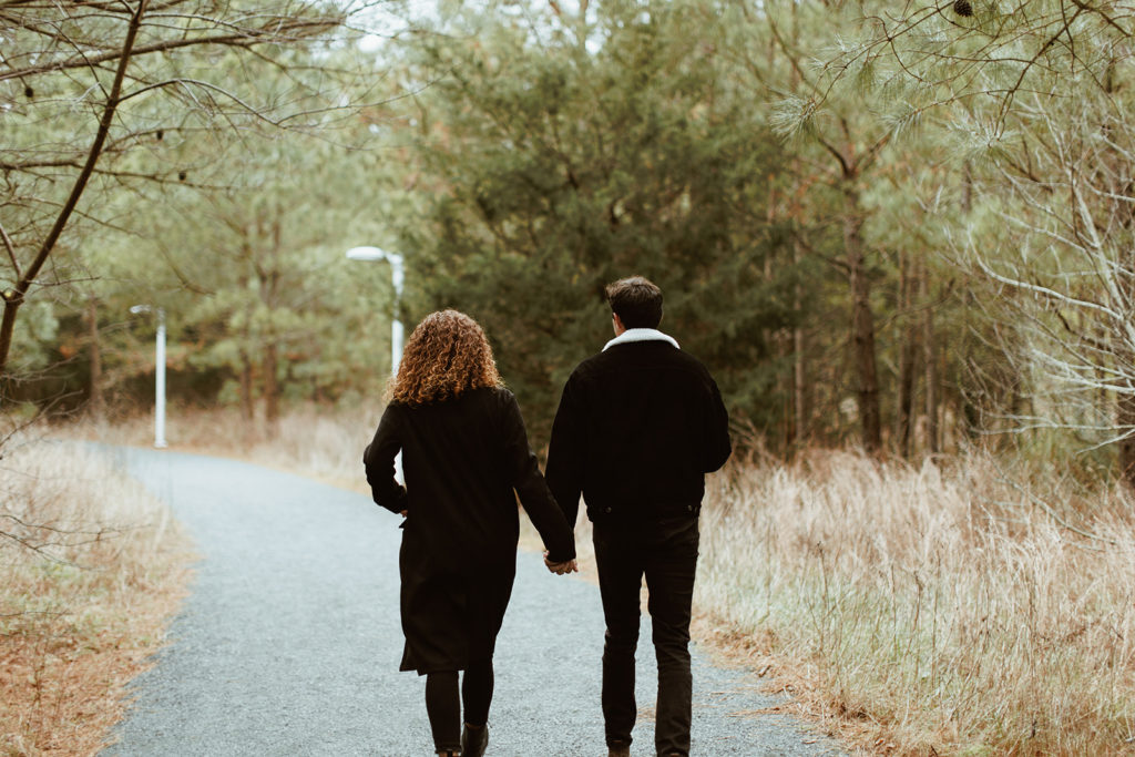 Couple holds hands walking through woods at engagement session taken by creative entrepreneur