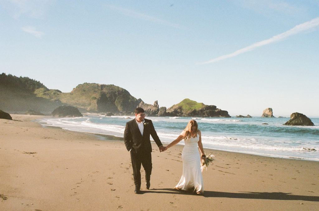 Couple walks along sand holding bouquet at beach elopement