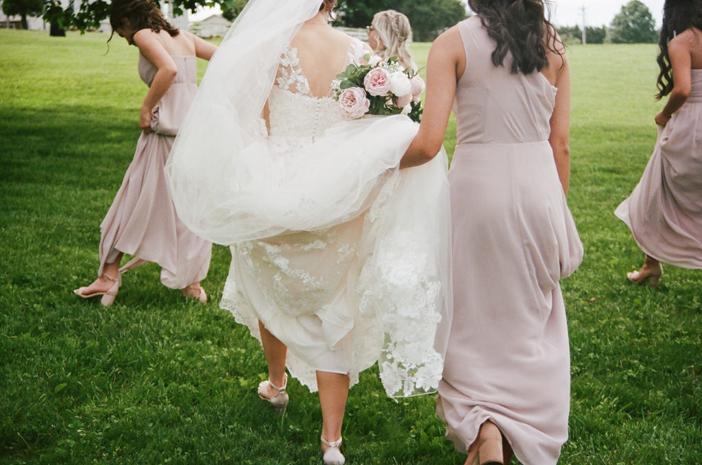 Bridesmaid holds bride's wedding dress as they walk with bridesmaids through field