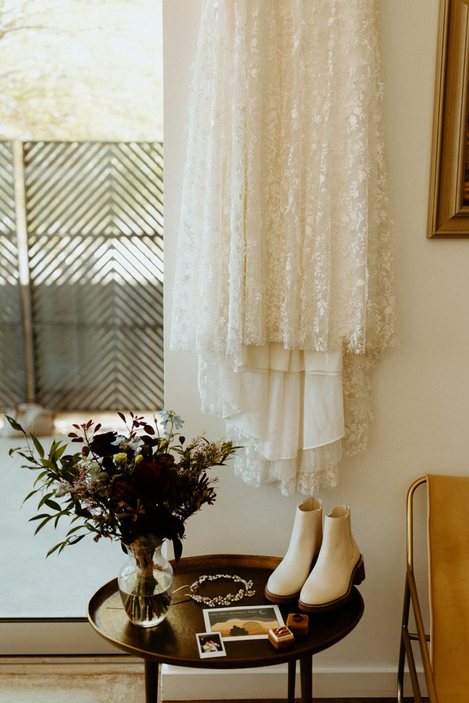 Wedding dress hangs over wedding shoes and bouquet