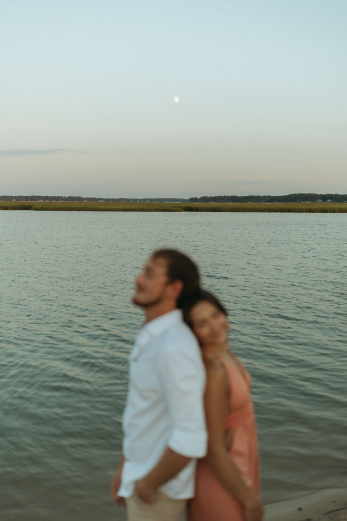 couple stands back to back beside water
