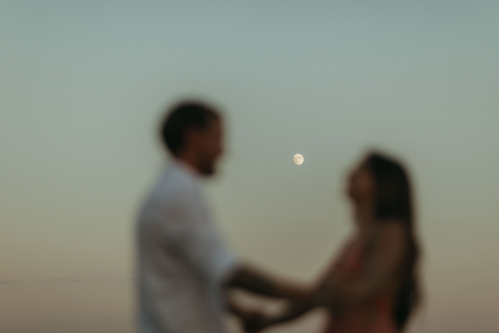 couple stands below moon at Virginia Beach sunset