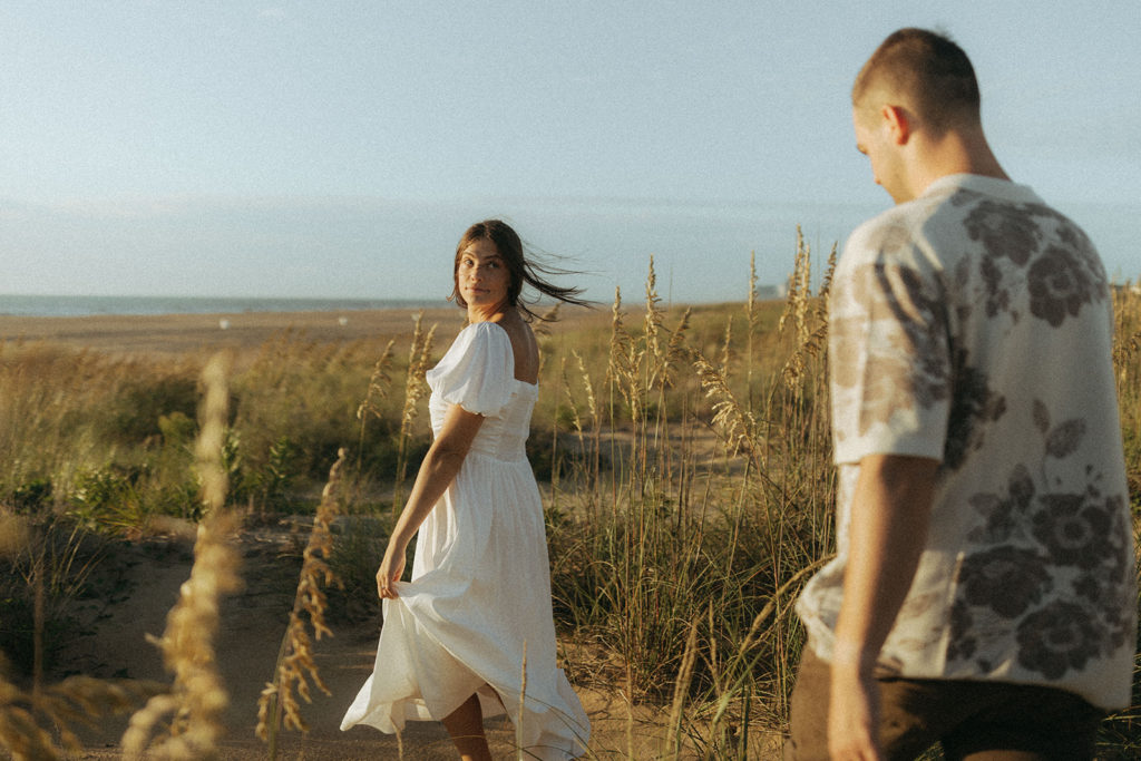 couple walks through grassy sand dunes at sunrise engagement session