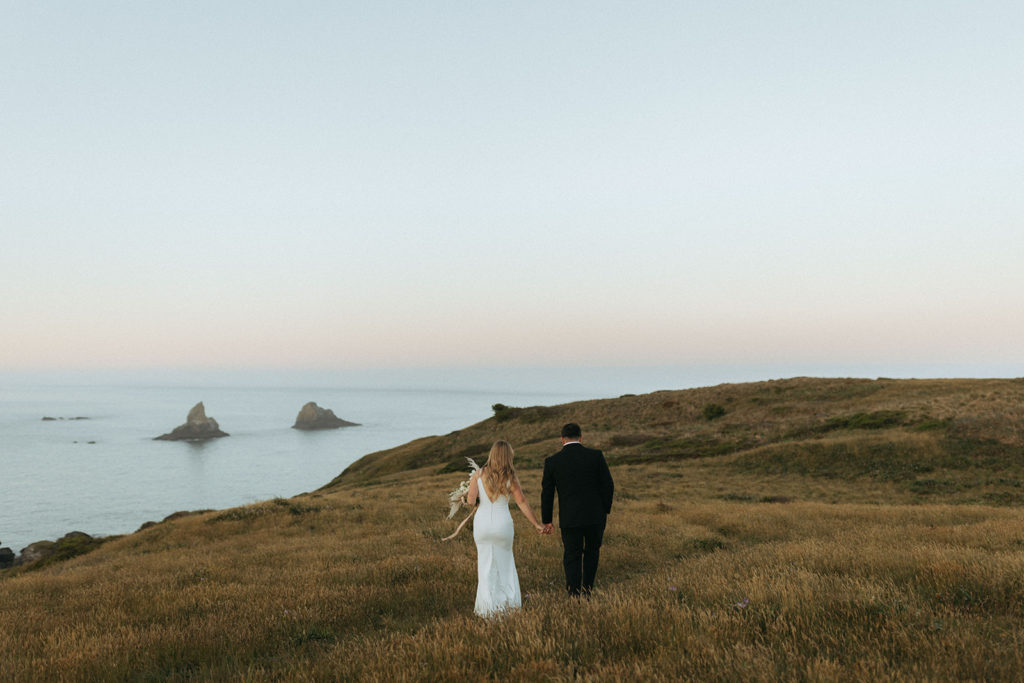 couple walks through field by water at Oregon coast wedding photoshoot