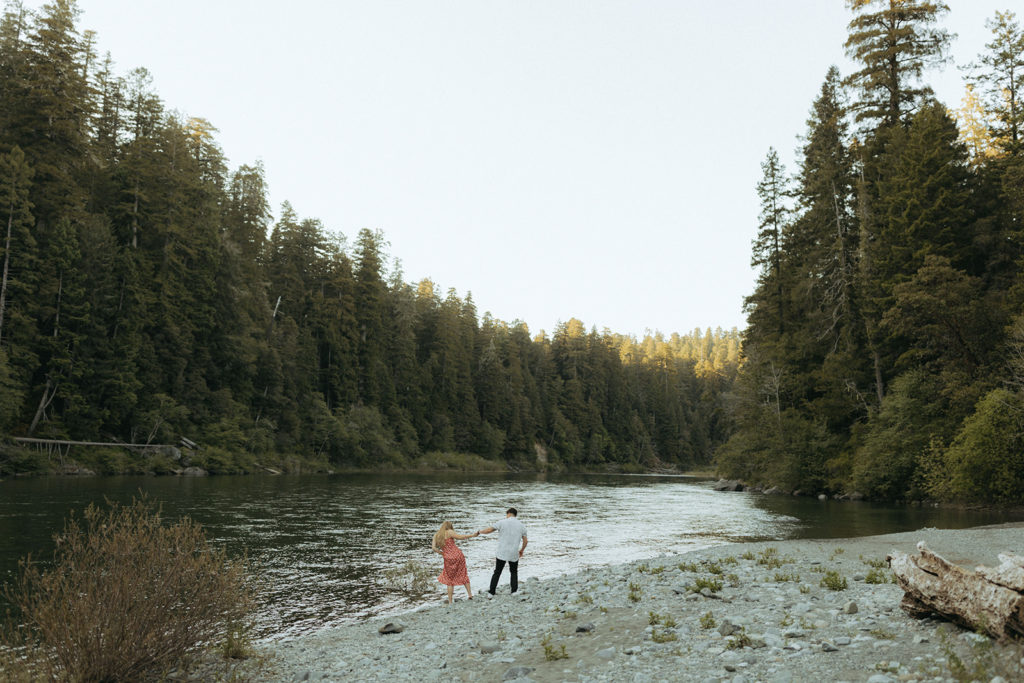 couple walks along river holding hands at Redwoods state park engagement photo session