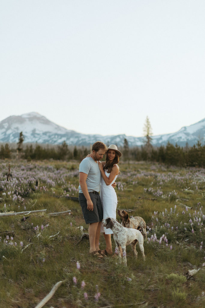 couple stand besides dogs at engagement session in field