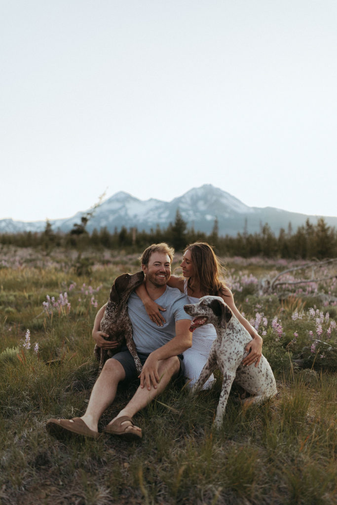 couple sits with dogs at engagement session in field
