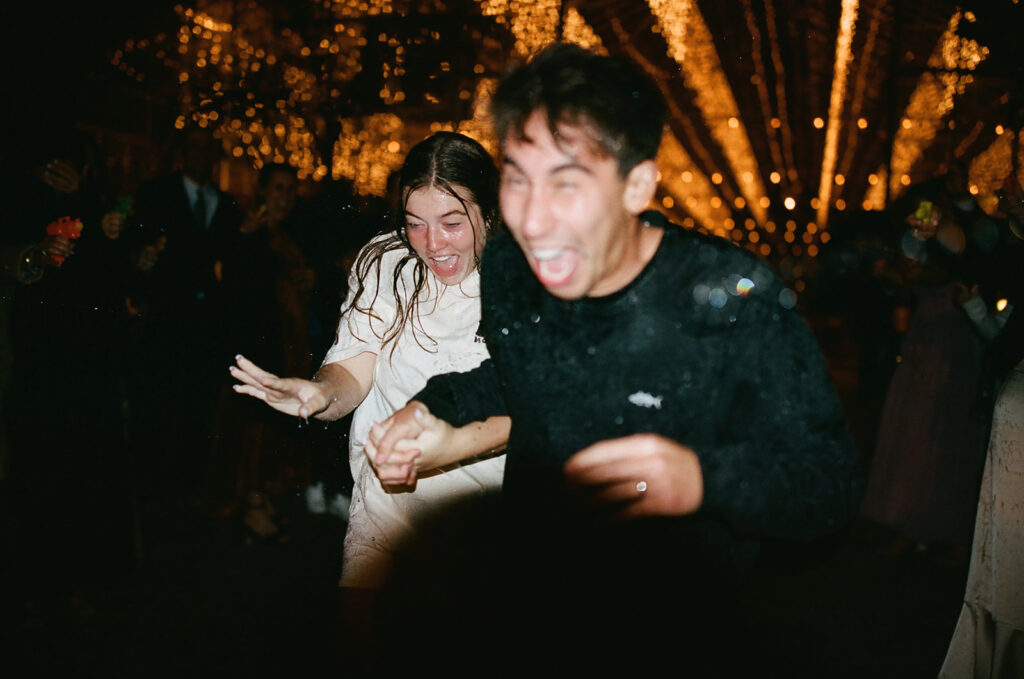 couple runs out of wedding exit hit by water guns as part of wedding film photography project
