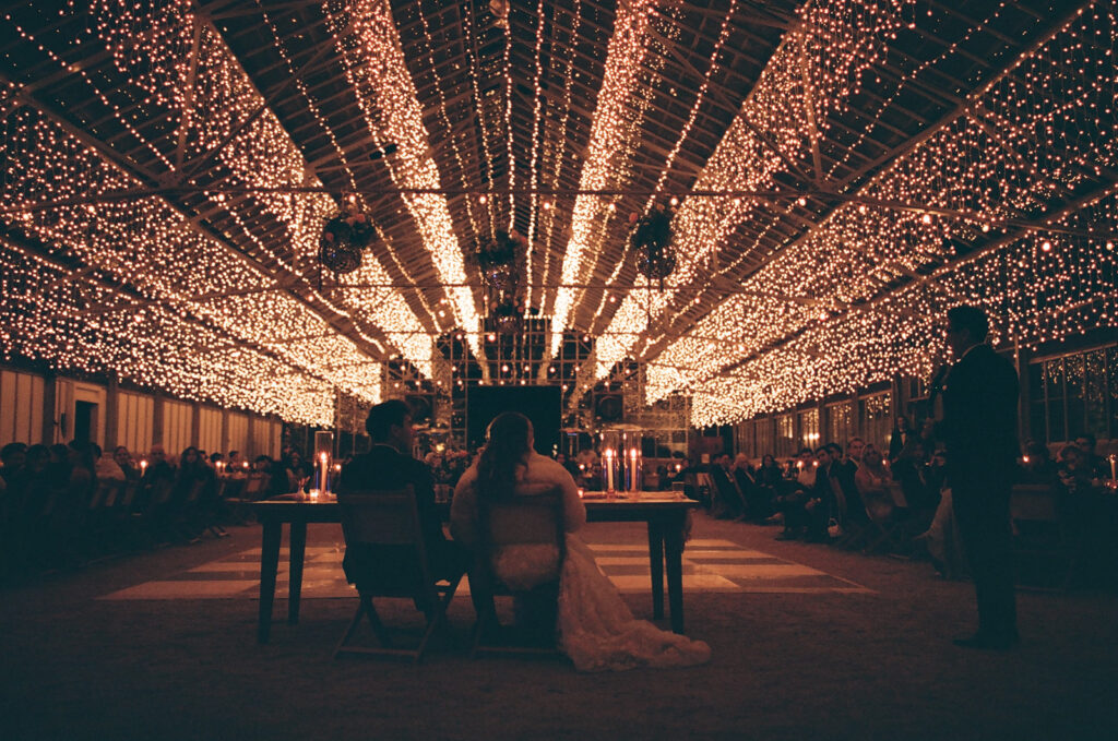 couple sits under twinkle-lights lit greenhouse as part of wedding film photography project