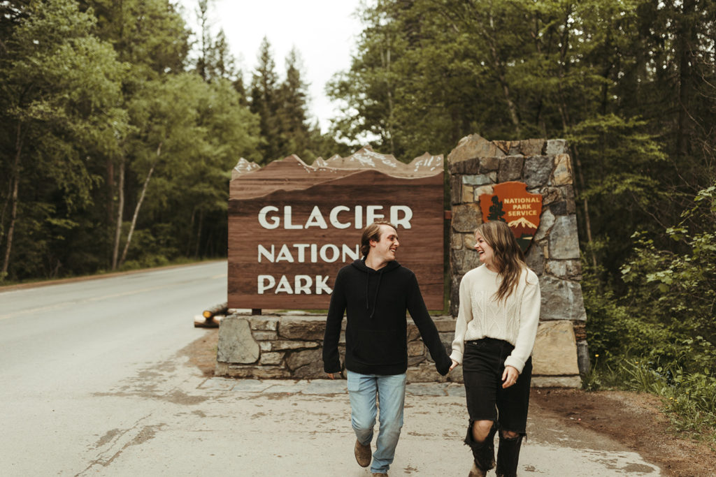 couple walks holding hands in front of Glacier National Park sign at engagement session