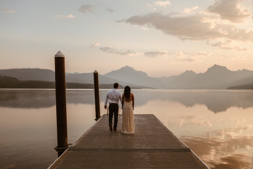 couple stands on pier at sunset lake in Glacier National park elopement