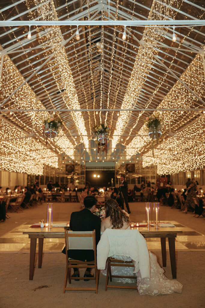 couple sits under twinkle lights during greenhouse wedding reception
