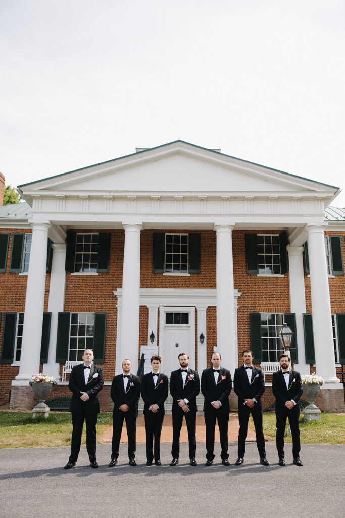 groom stands surrounded by wedding party in front of historic house