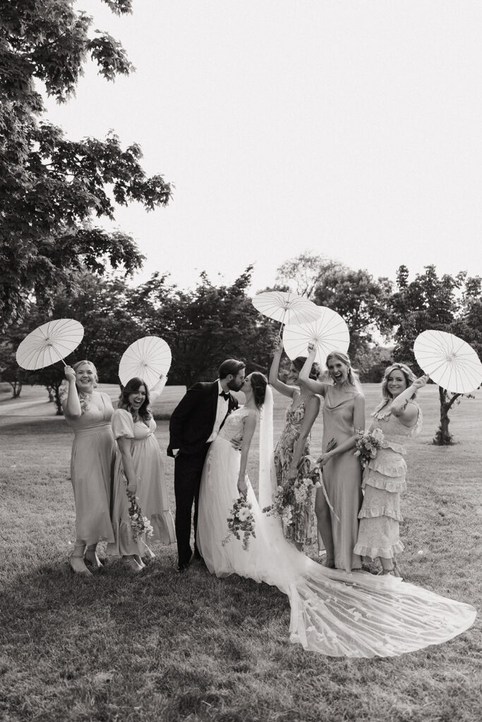 couple kisses beside bridesmaids cheering holding umbrellas 