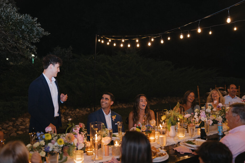 couple sits with at long table under twinkle lights at wedding reception in backyard