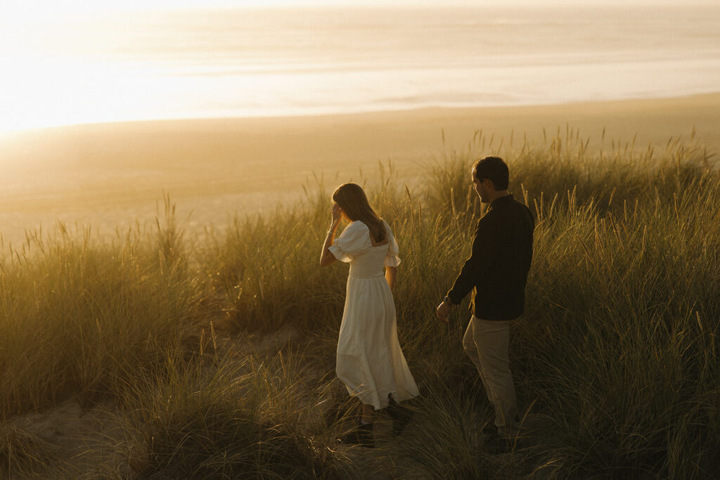 couple walks through high grass at Cannon Beach sunset engagement session