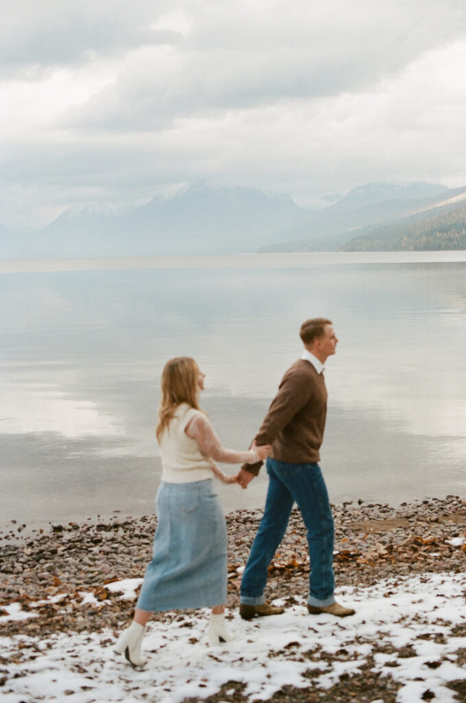 couple walks holding hands at snow-covered lake at Glacier National Park engagement